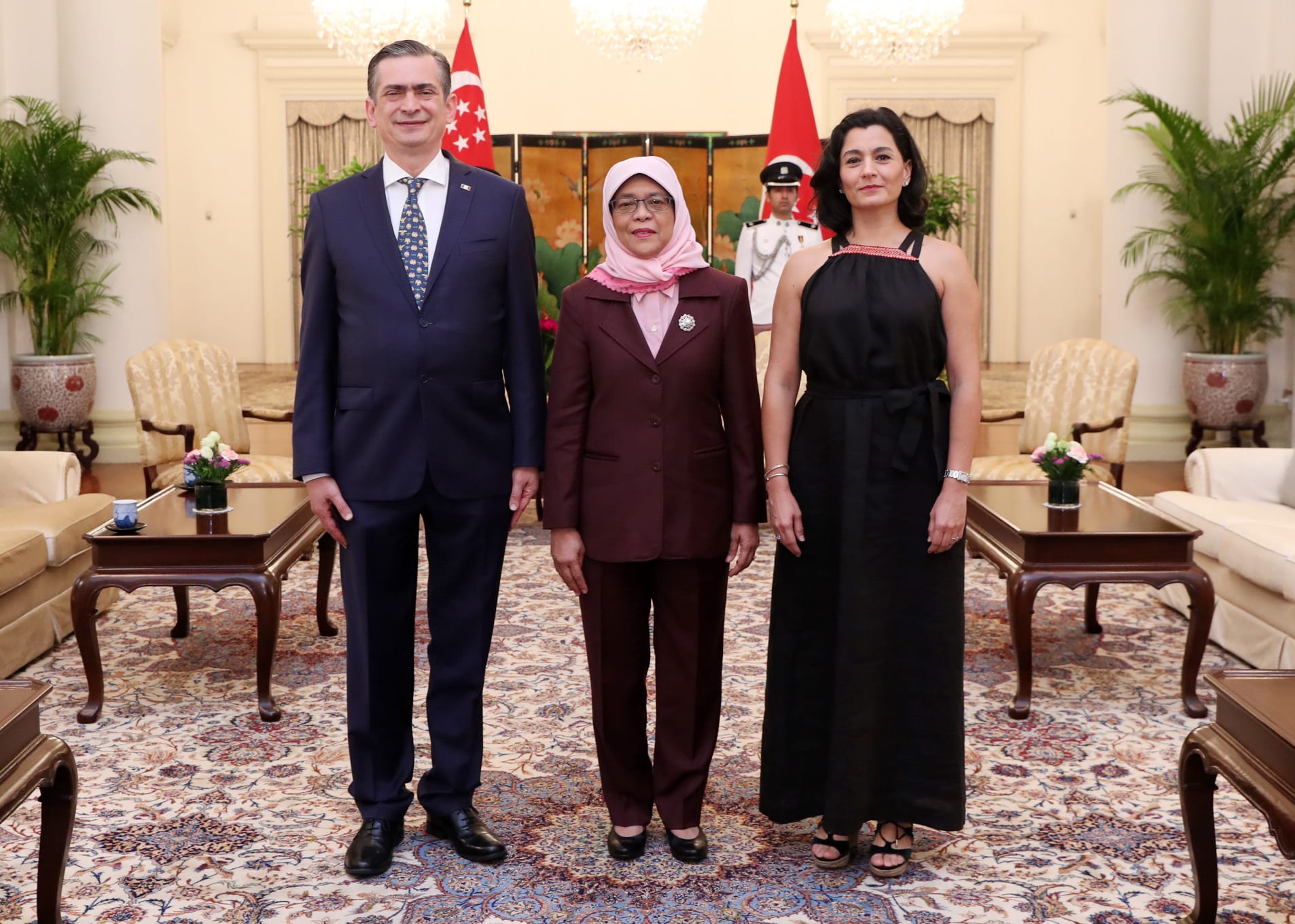 Three people posing indoors in front of flags and a guard, with ornate decor.